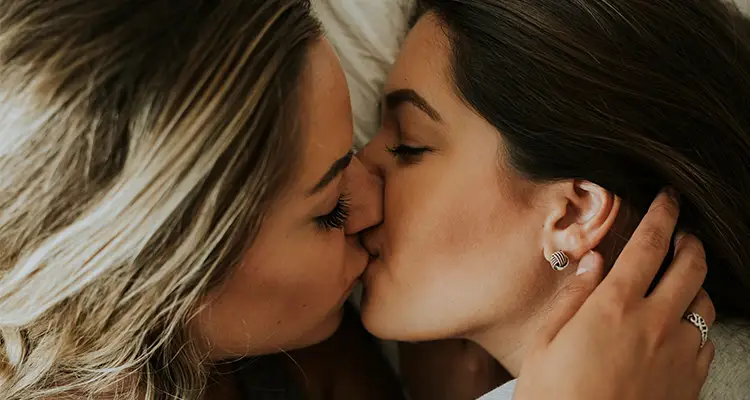 Blonde woman and brunette woman lying on a white pillow kissing each other on the lips.