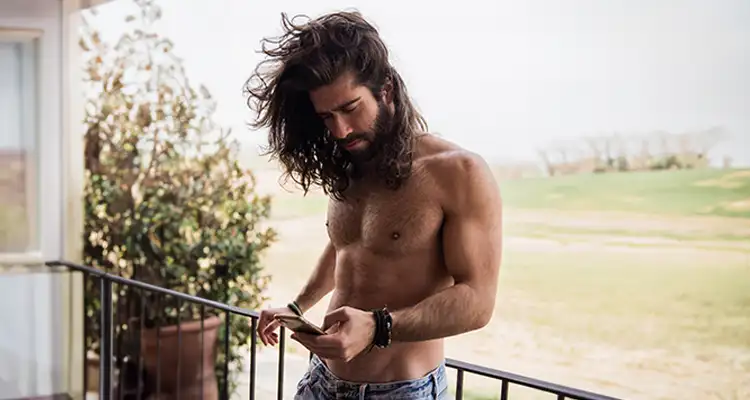 Long-haired brunette man, shirtless and leaning against the railing of a veranda with a green field in the background.