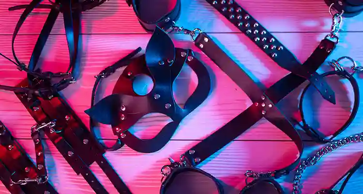 Black leather bondage gear including a mask, whips and collars on a wooden floor under red and blue neon lights.