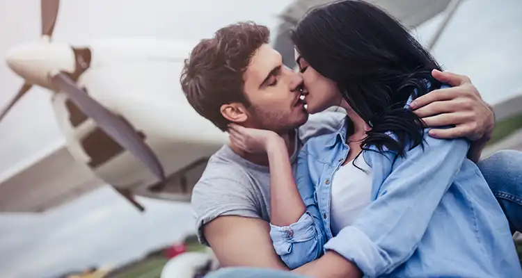 Brunette woman in white shirt and blue jacket and man in grey shirt kissing with airplane behind them.