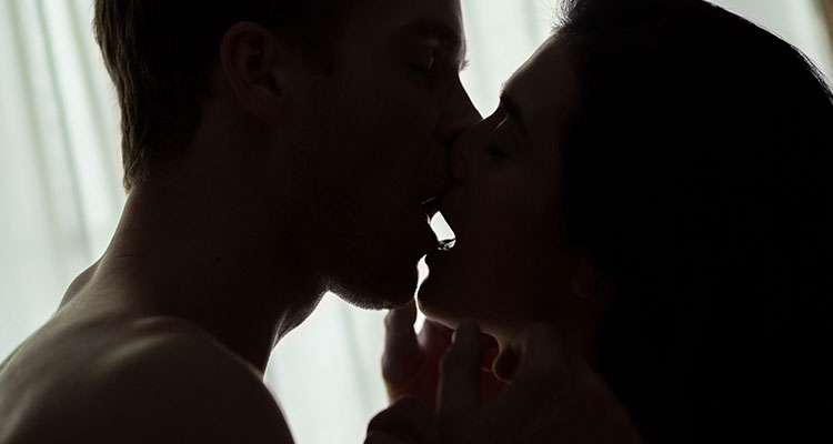 Brunette man and woman in badly lit room kissing passionately with their eyes closed. White curtains in the background.