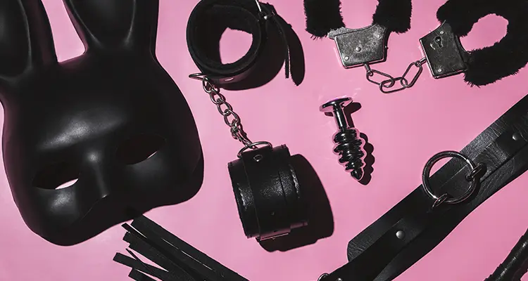 Many black and silver BDSM sex toys including a bunny mask lying on top of a light pink background.