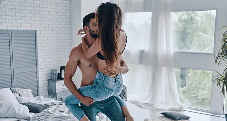 Brunette shirtless man lifting a brunette woman in black bra and jeans while she wraps her hands around his neck.
