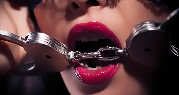 Half-parted lips of a woman bringing the cain of her handcuffs up to her mouth to bite down on them.