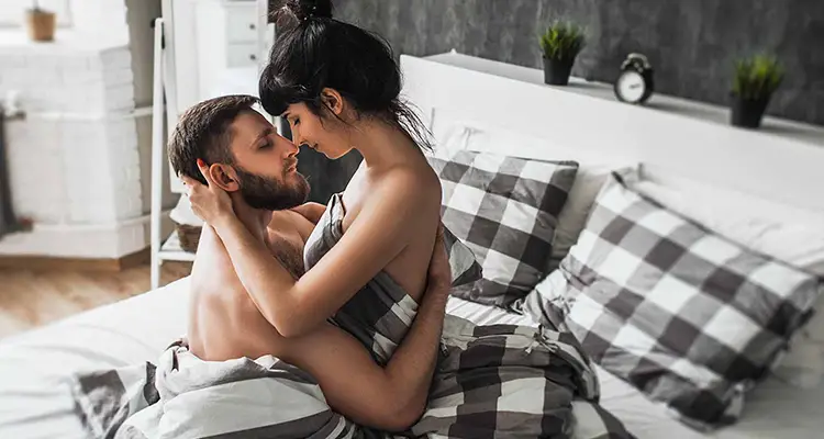 Brunette man and woman naked under black and white checkered sheets hugging each other close.