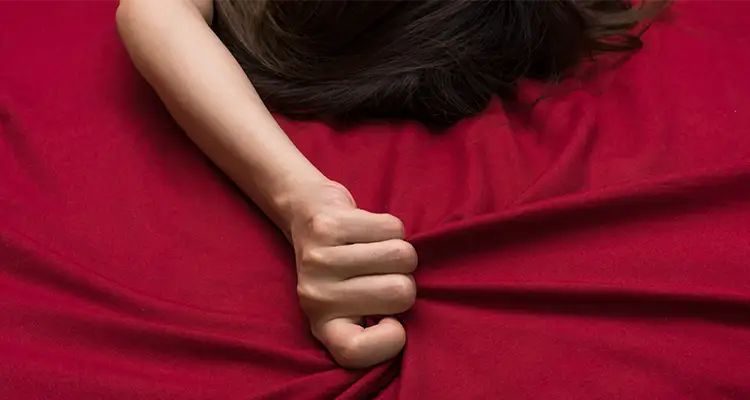 Brunette woman lying in a bed of red sheets, clenching the sheets with her palm on top of her head.