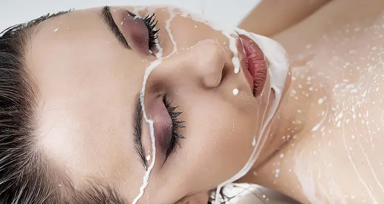 Brunette naked woman wearing purple eye shadow with her face and chest covered in white liquid.