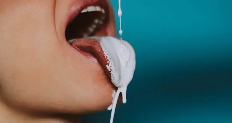 Opened mouth of a woman with her tongue out while white liquid is being dripped down onto her tongue.