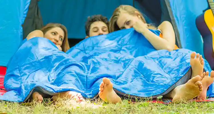 Naked man with two women covered in blue sheets as they lie on grass with a camping tent behind them.