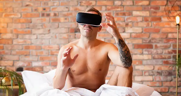 Blonde shirtless man in bed covered by white sheets wearing a VR headset with his hands in front of him grabbing the air.