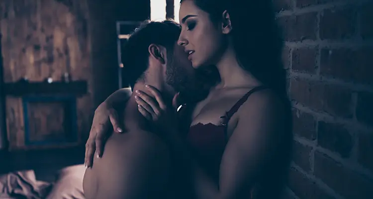 Brunette man holding woman up and against the wall while kissing her neck. He is shirtless and she is in black lingerie.