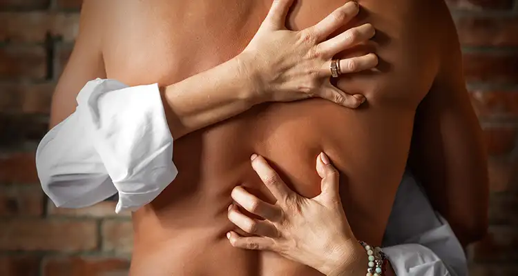 Shirtless man hugging a woman in white shirt who is digging her nails in his back. Brick wall in the background.