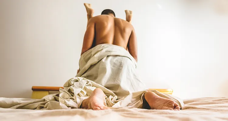 Shirtless man covered by white bedsheets leaning down to a woman whose feet rest on each of his shoulders.