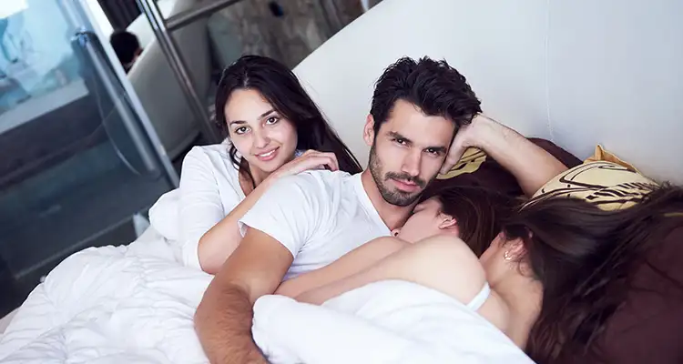 Brunette man in bed with three brunette woman covered in white sheets, looking at the camera with a smug look.