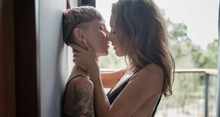 Blonde woman in black lace bralette holding the face of a short-haired woman against the wall and leaning in to kiss her.