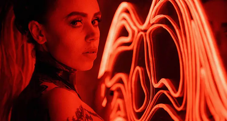 Woman wearing black choker with multiple tattoos on her body looking at the camera, standing in a room lit in red.