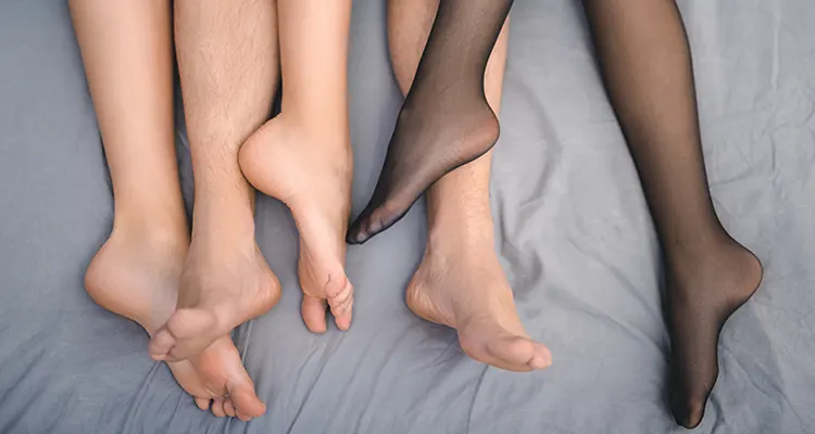 Several pairs of feet peaking through from under grey sheets clearly lying on a bed and intertwining in between each other.