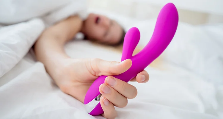 Woman lying in bed while moaning, holding a pink dildo with her body covered in white sheets.