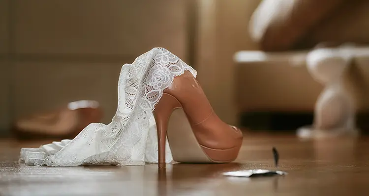 Beige high heel on the ground with lace white panties hanging off the side of it with an opened condom wrapper next to it.