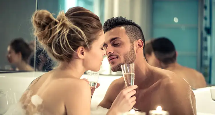 Blonde woman and brunette man holding champaign glasses in bathtub full of bubbles, leaning in to kiss each other.