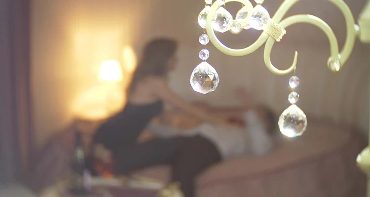 Chandelier in focus with blurred woman in the background pushing a man on the bed next to alcohol bottles.