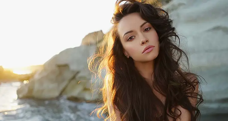 Brunette attractive woman with her hair covering her chest in front of a cliff by the sea.