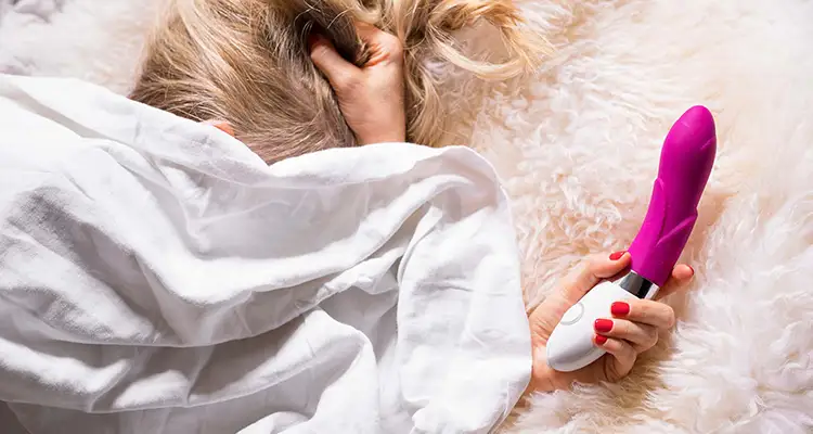 Blonde woman under white bed sheets lying on fluffy blanket holding her head and a pink vibrator.