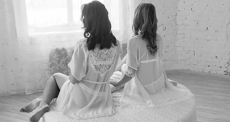 Two women in see-through lacy lingerie, sitting next to each other holding hands and looking out the window in front of them.