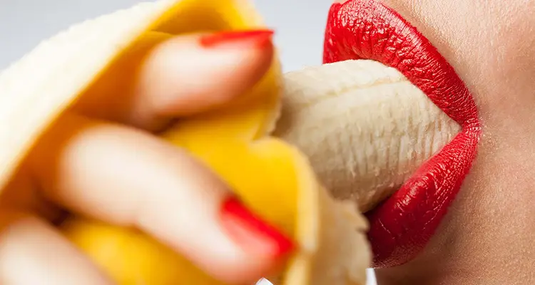 Mouth of a woman with red lipstick sensually biting down on a half-peeled banana. Her nails are coloured red.