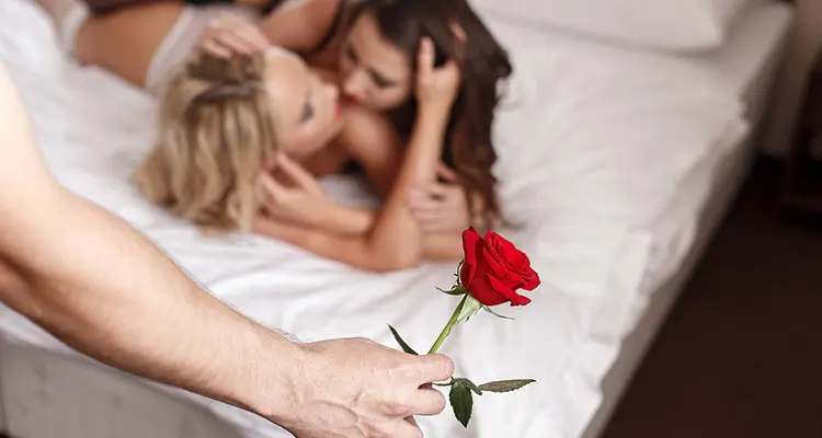 Brunette and blonde girls kissing in their lingerie on a bed and hand of a man holding red rose approaching them.