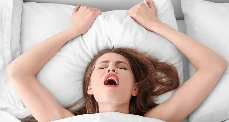 Brunette woman in bed holding the pillow behind her, moaning in ecstasy. White sheets cover her body.