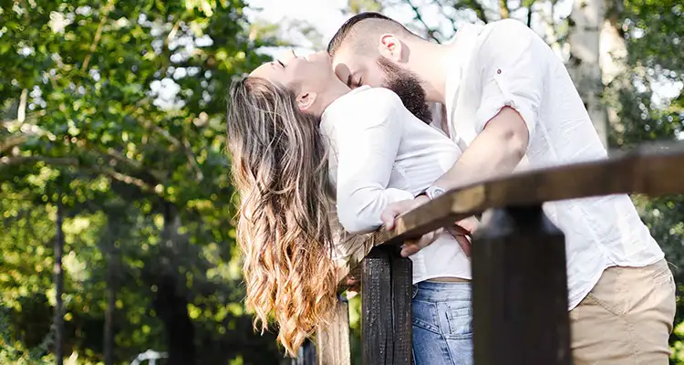 Woman leaning against a wood railing getting kissed on the chest by a bearded man pushing her against the railing.