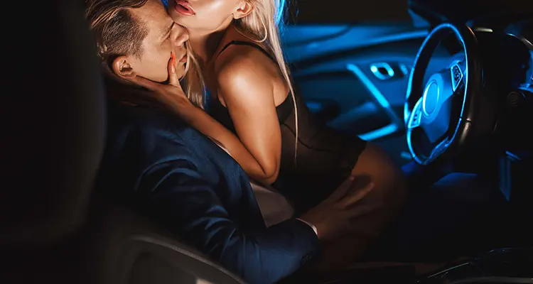 Blonde woman in black lace lingerie straddling a man wearing a suit sitting in the driver's seat of a car.