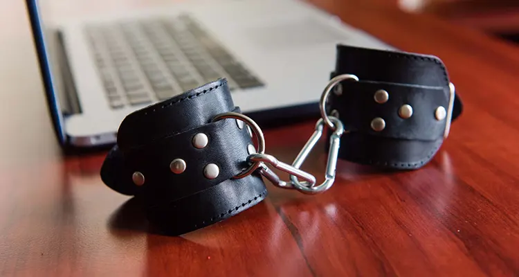 black leather handcuffs connected by chains on wooden desk with grey laptop in the background.