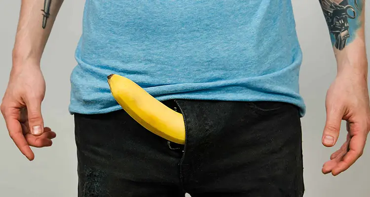 Man wearing black jeans has a banana sticking out from his zipper. He is wearing a light blue shirt and has colorful tattoos on his arms