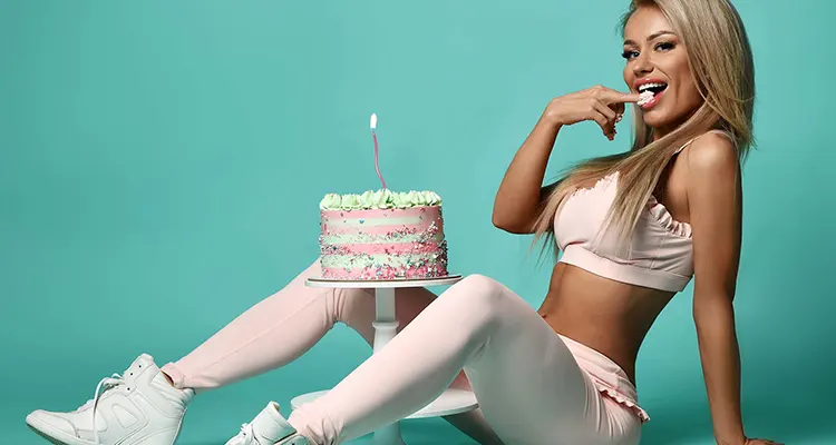 Blonde older woman wearing light pink trousers and crop top with cake between her legs, licking her fingers.