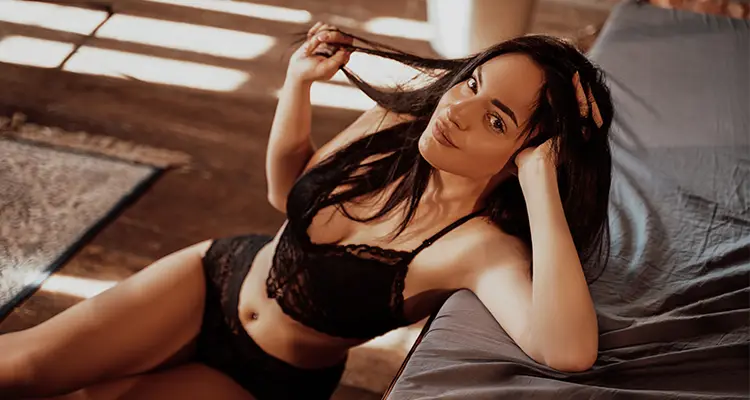 Brunette woman in lace black lingerie sitting on the ground and supporting her head with her hand. Her elbow is on the bed.